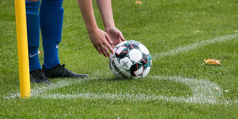 El corner de un campo de fútbol, una pelota sobre el césped y unas piernas preparándose para chutar. 