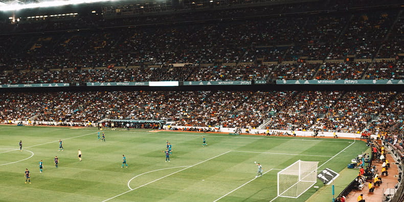 Vista de un estadio de fútbol con jugadores en acción sobre el campo y una multitud de espectadores observando del partido desde las gradas.
