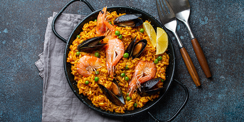 Plato de paella en una paellera con pollo, judías verdes y trozos de limón como decoración, sobre una mesa con mantel. 